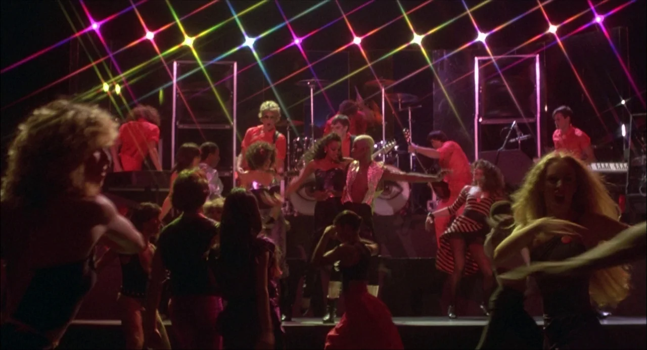 Xanadu (1980) movie still: wide — A dark concert stage is filled with performers in 1980s clothing and colorful lasers. The band plays instruments…