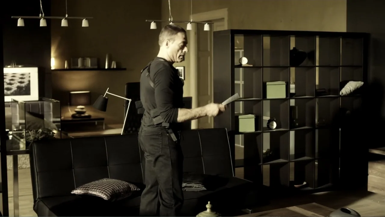 Assassination Games (2011) movie still: medium — A modern living room features a dark black bookshelf and a low sofa. A man in a black tactical…
