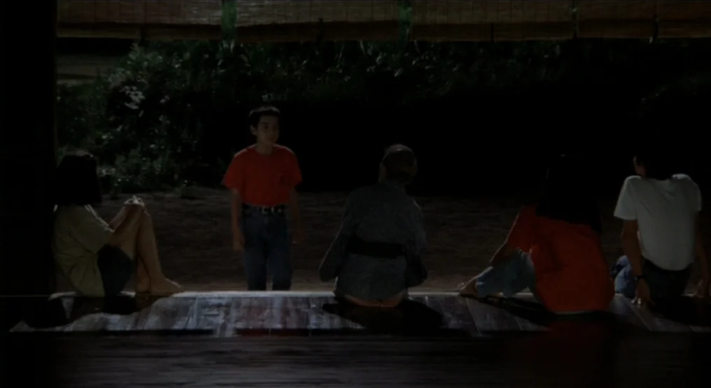 Rhapsody in August (1991) movie still: wide — A dark wooden Japanese-style house veranda overlooks a shaded, dark outdoor area at night. Five…