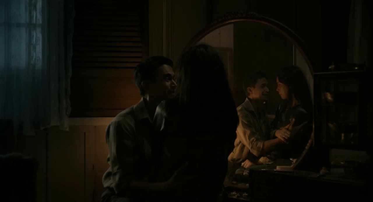 In My Mother’s Skin (2023) movie still: medium — A dimly lit space features a wooden vanity with a large circular mirror. A couple embraces while…
