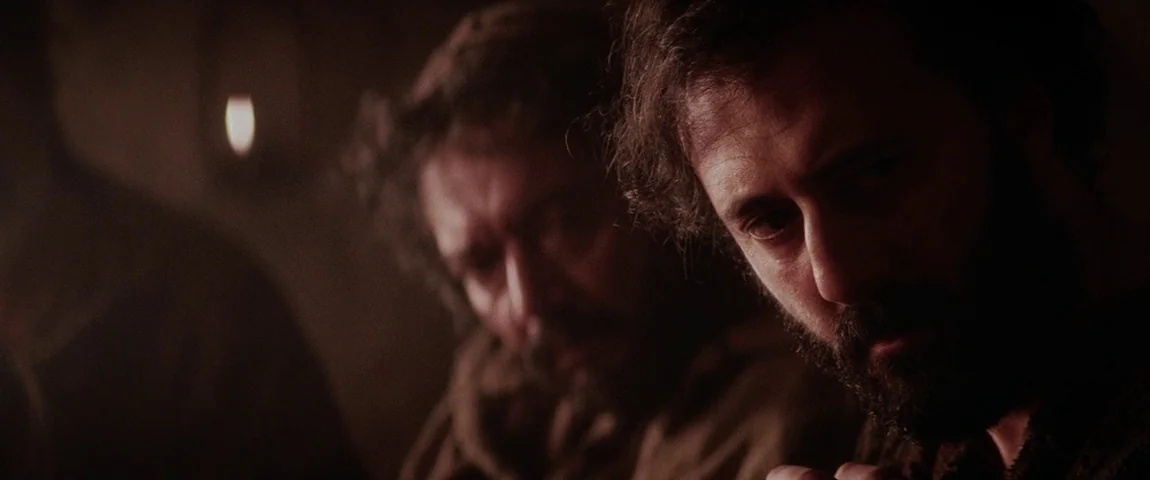 The Passion of the Christ (2004) movie still: closeup — A dark, dimly lit stone interior environment with minimal visible features. Two men with…