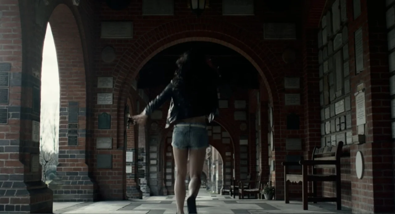 Back to Black (2024) movie still: fullbody — A stone and brick corridor features arched openings and walls covered in small plaques. A young woman…