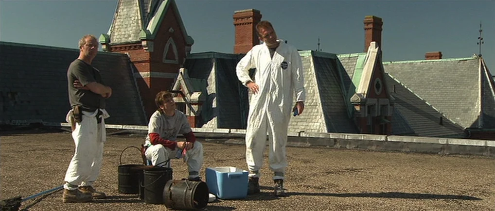 Session 9 (2001) movie still: fullbody — A flat rooftop covered in gravel features several black buckets and a blue cooler. Three men wearing work…