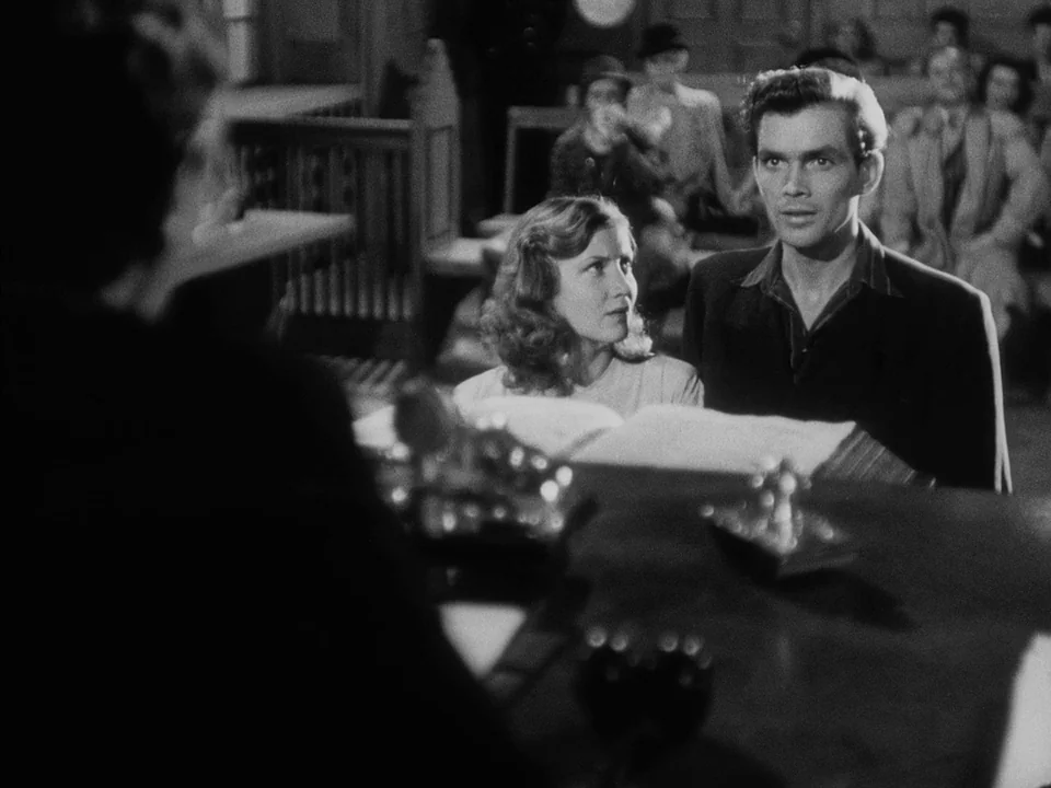 It Rains On Our Love (1946) movie still: medium — A courtroom with a wooden witness stand and spectators blurred in the background. A man and woman…