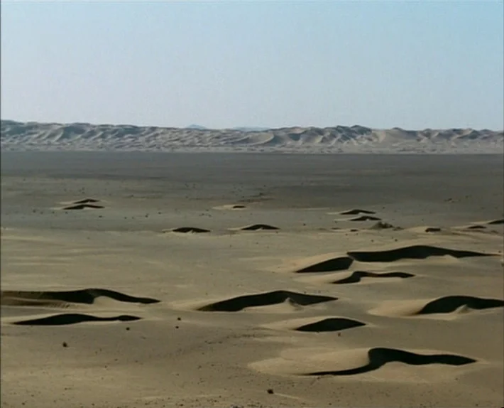 Fata Morgana (1971) movie still: establishing — A vast, arid desert landscape features rolling sand dunes under a clear blue sky. The flat horizon…