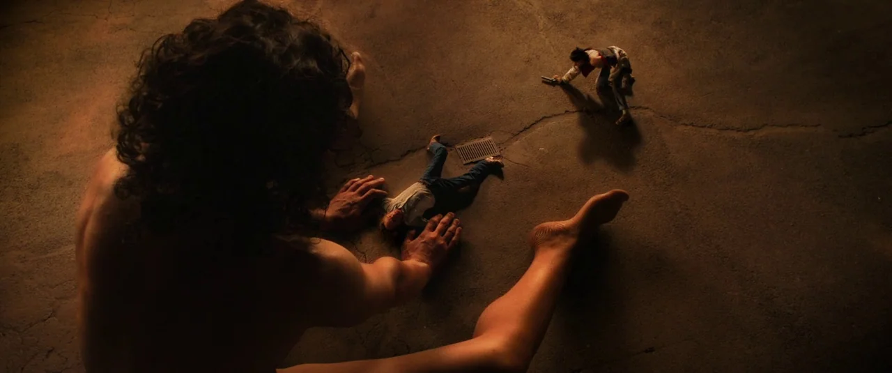 Love Lies Bleeding (2024) movie still: wide — A flat concrete surface at night with cracks and a central metal drainage grate. A giant figure towers…