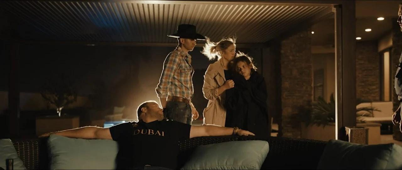 American Sicario (2021) movie still: fullbody — A modern outdoor patio at night features a sofa under a covered roof structure. Four people stand or…