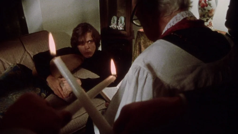 Martin (1977) movie still: medium — A dark living room contains a couch and decorative religious or vintage ornaments. A man holds two candles…