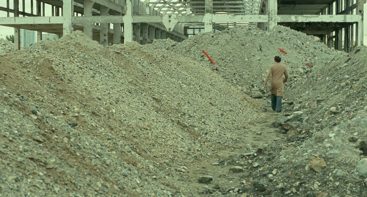 Saint Laurent (2014) movie still: wide — An abandoned industrial structure looms over a field of gravel and debris. A man in a light coat walks…