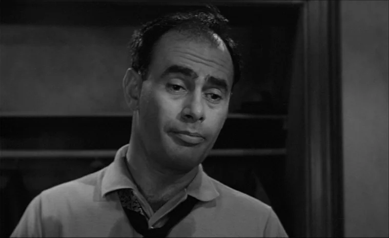 12 Angry Men (1957) movie still: closeup — An interior space with shelves visible in the background indicating storage. A middle-aged man stands in…