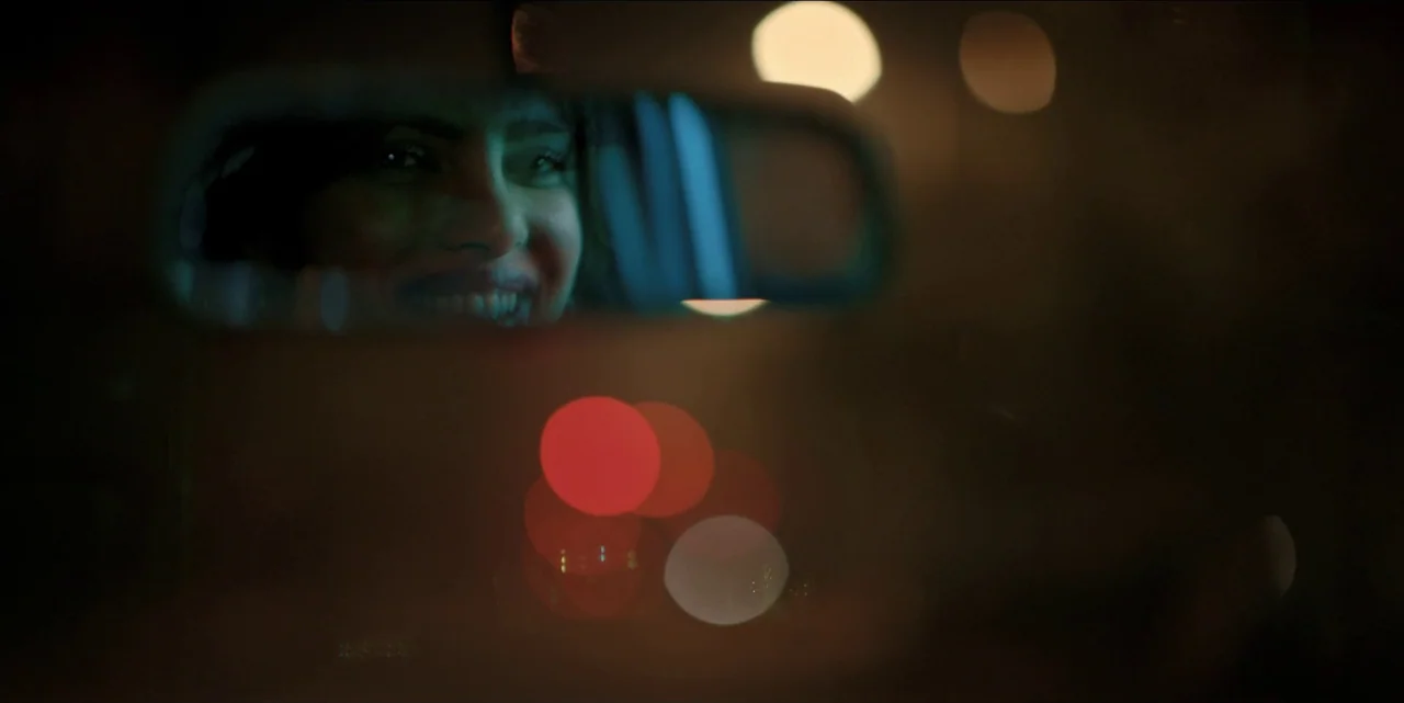 The White Tiger (2021) movie still: closeup — A dark vehicle interior with a view through a rearview mirror at night. A woman appears in the…