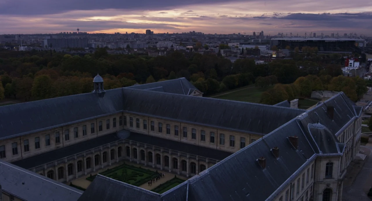 Zombi Child (2019) movie still: establishing — A large stone building with a central garden courtyard overlooks a distant city skyline. The twilight…