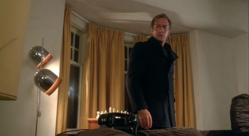 The Silent Partner (1978) movie still: medium — A living room interior features a floor lamp, mustard-yellow curtains, and a chessboard. A man in a…