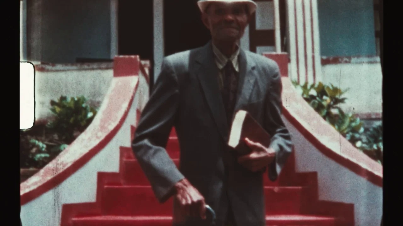 Black Mother (2018) movie still: fullbody — A man descends stairs leading to a building with red-painted treads and white railings. He carries a…