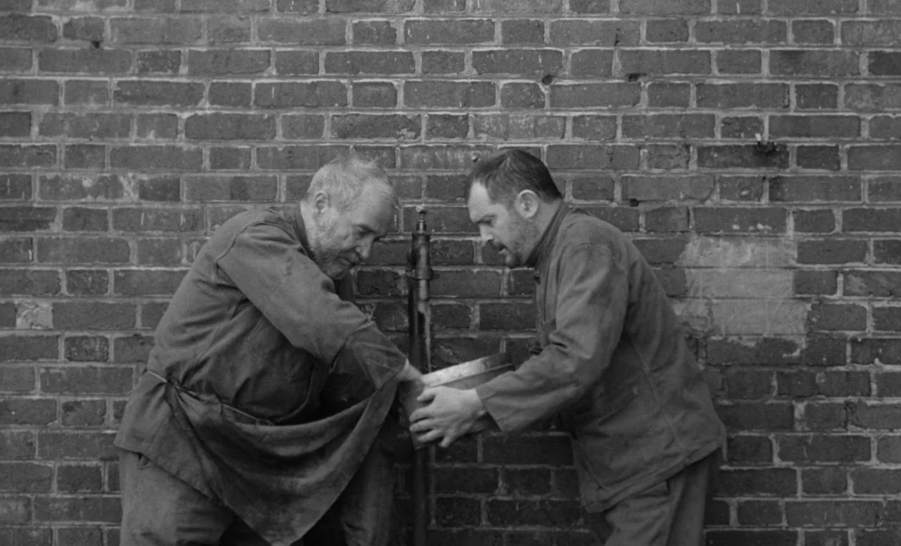 Goto, Island of Love (1969) movie still: medium — A dark brick wall provides a textured backdrop for an outdoor manual water station. Two men in…
