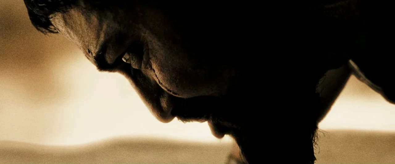 300 (2006) movie still: closeup — A man in a desert environment bows his head low in extreme bright sunlight. The camera focuses on the sharp…