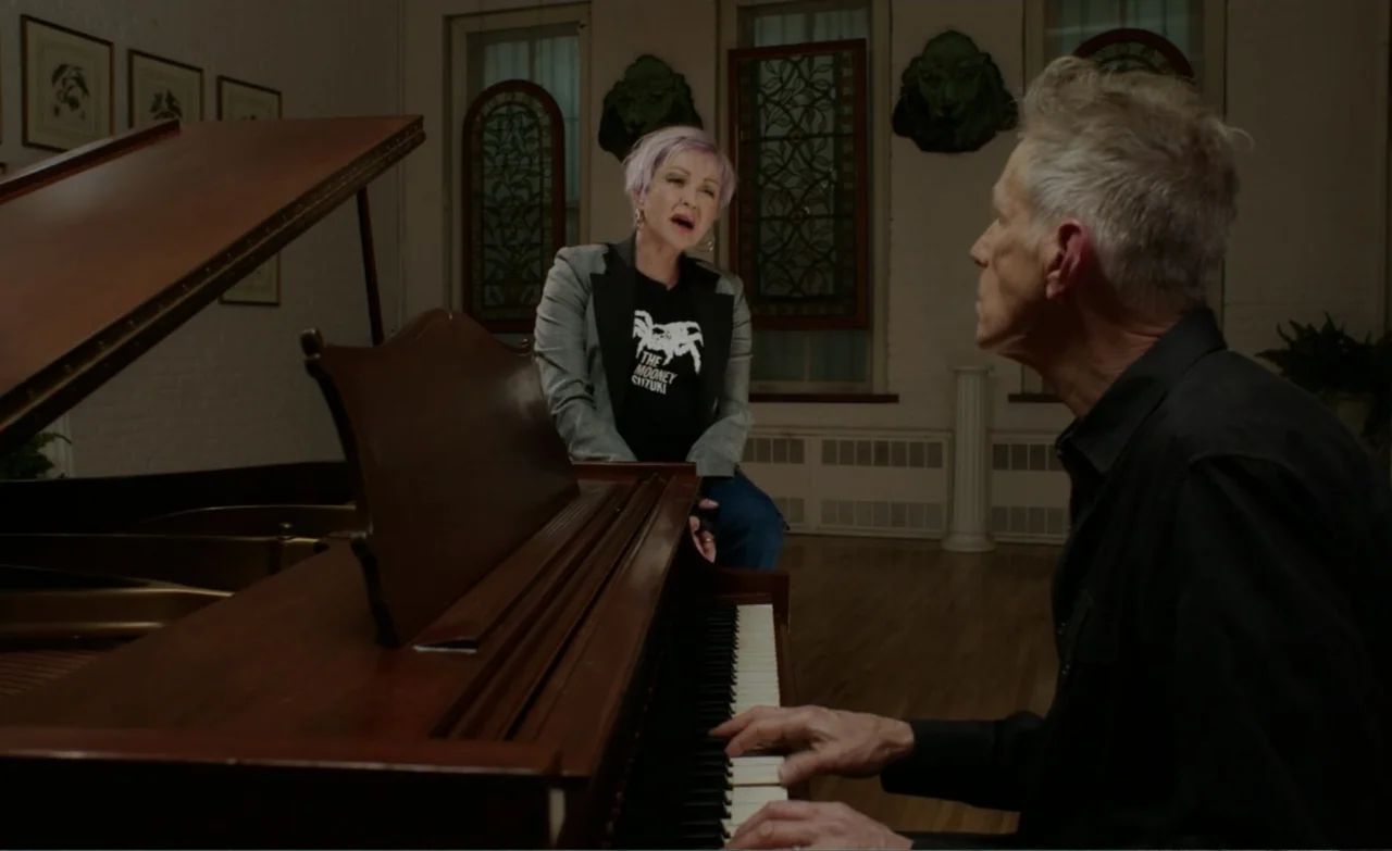 Let the Canary Sing (2023) movie still: medium — A room with wooden flooring features a large piano and tall arched windows. A man plays the keys…