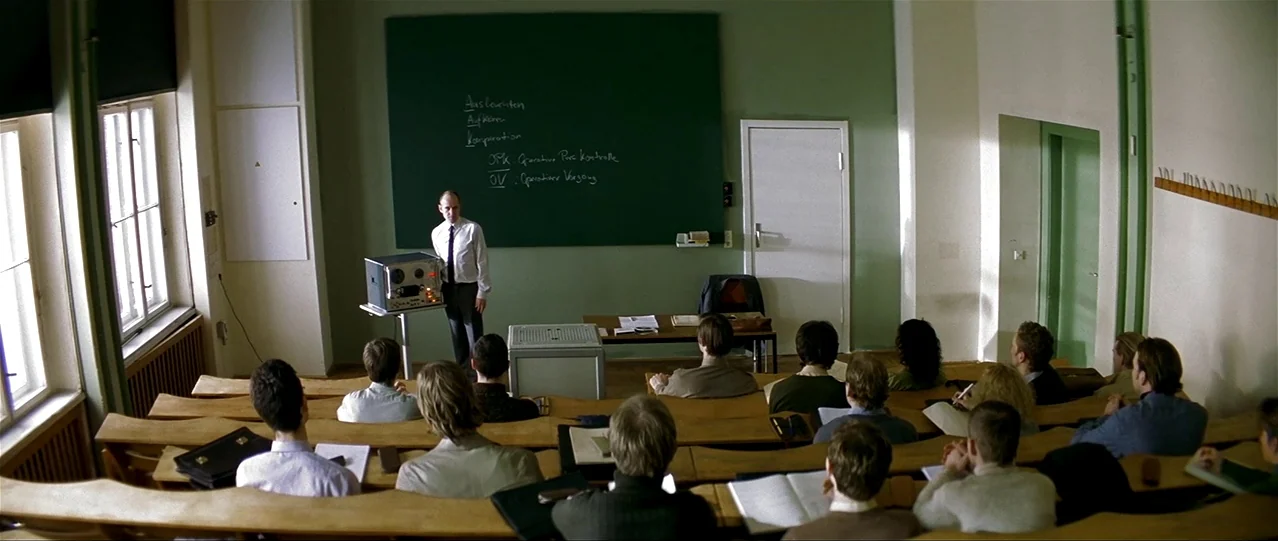 The Lives of Others (2006) movie still: wide — A tiered lecture hall with green walls and wooden desks faces a green chalkboard. A man in a dress…