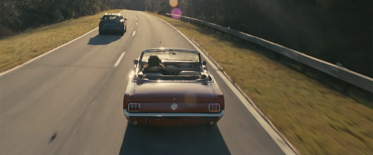 Companion (2025) movie still: wide — A paved highway curves through an area with dry grassy hills and metal railings. A red vintage convertible…