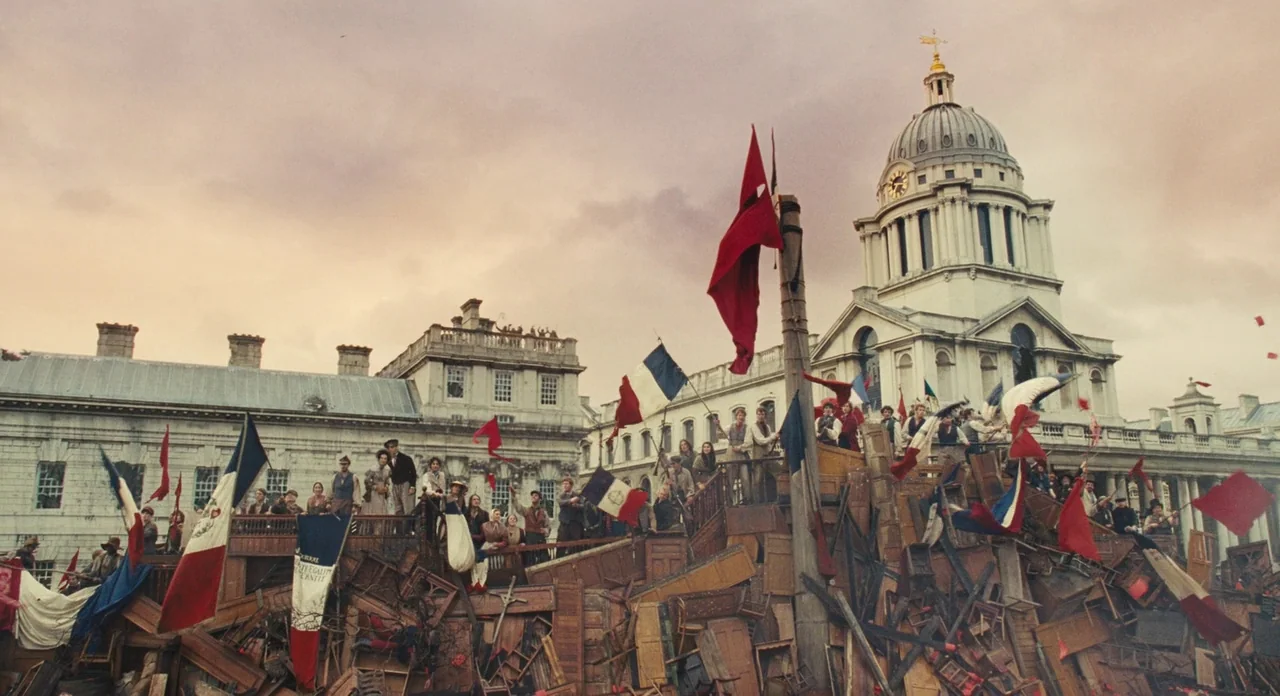 Les Misérables (2012) movie still: wide — A sprawling barricade constructed from discarded wooden furniture sits before a classical stone building.…
