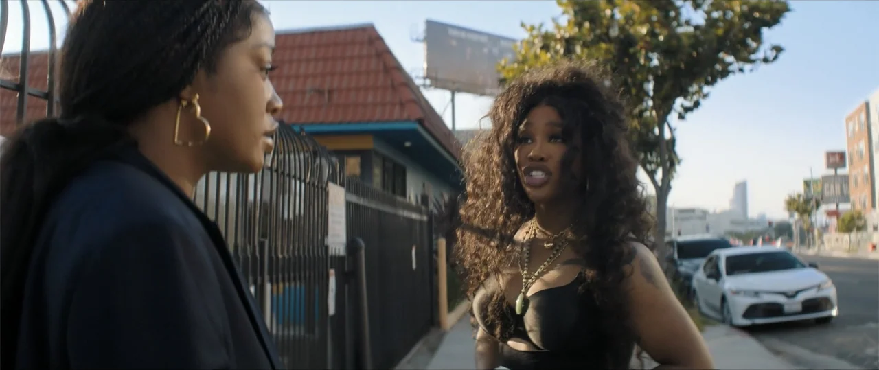 One of Them Days (2025) movie still: medium — A sidewalk borders a black metal fence with a commercial building in the background. Two women face…