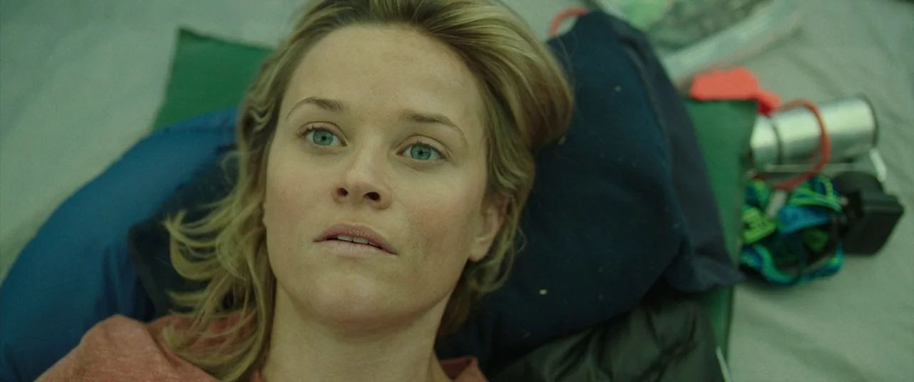 Wild (2014) movie still: closeup — The interior of a cramped tent is dimly lit with a green-toned ambient light. A woman lies on her back on a mat,…