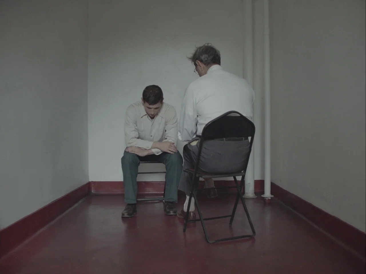 The Mountain (2018) movie still: fullbody — A stark room features plain white walls and a deep red floor molding. Two men sit on metal folding…