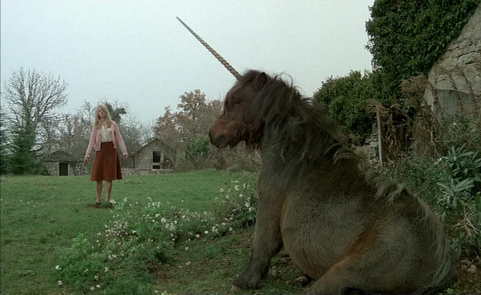Black Moon (1975) movie still: fullbody — A green pasture features a small stone outbuilding and a rock wall backdrop. A blonde woman stands at a…
