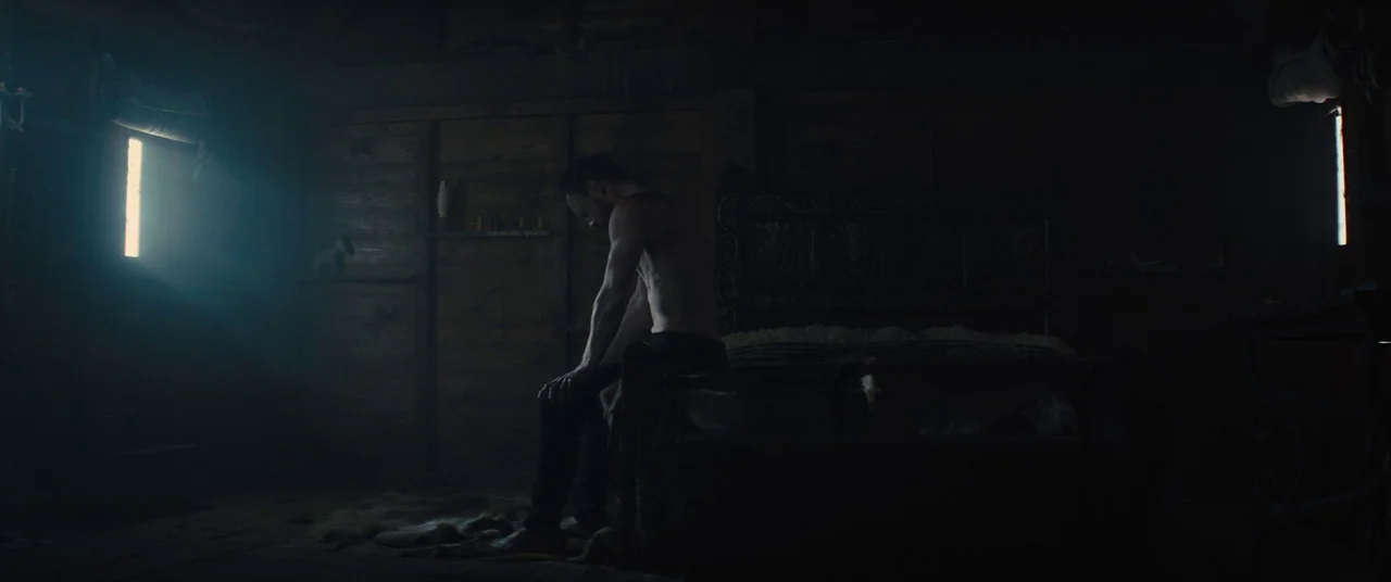 Macbeth (2015) movie still: medium — A dimly lit timber room features dark wooden walls and an old bed frame. A man sits shirtless on the edge of…