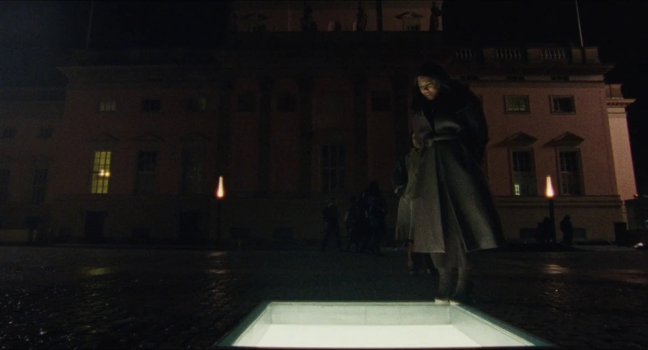 Origin (2023) movie still: fullbody — A dark, paved plaza at night features a large, illuminated glass panel inlay. A woman stands over the light…