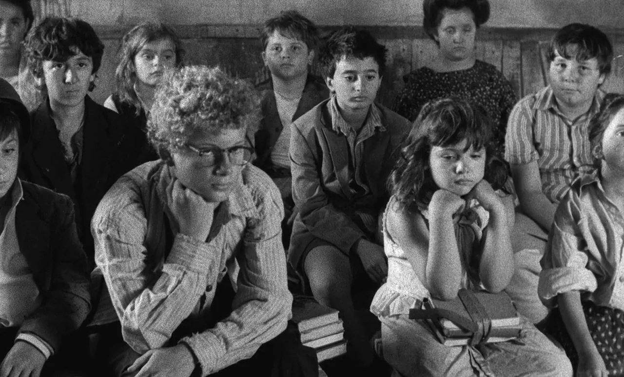 Go Down Death movie still: medium — A crowded schoolroom with children sitting in wooden benches arranged in rows. The children face forward with…