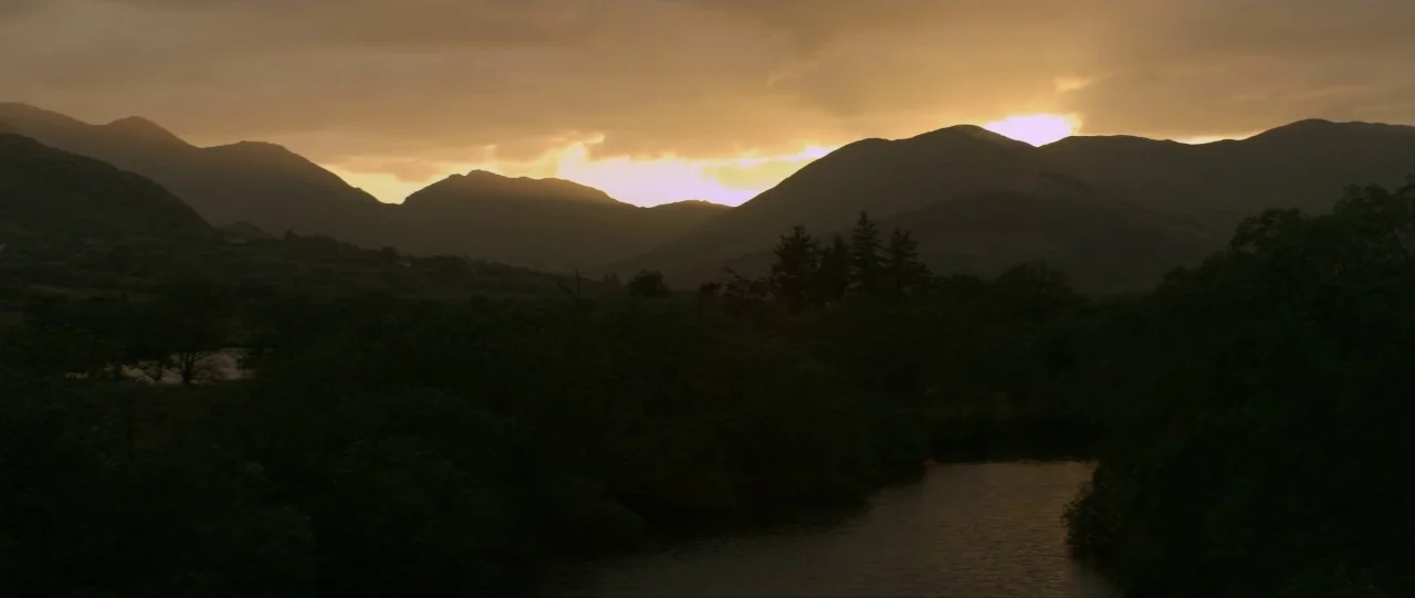 Silence (2012) movie still: establishing — A dark, silhouetted valley sits below a range of mountains at sunset. Golden light breaks through a layer…