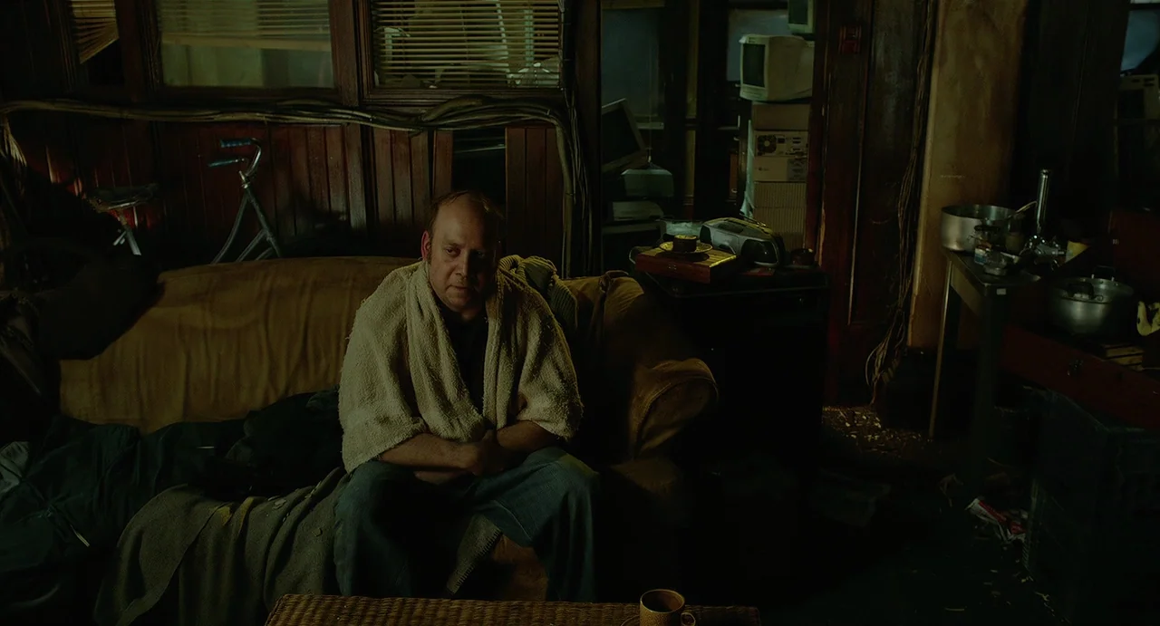 Cosmopolis (2012) movie still: medium — A cluttered apartment space features dark wood paneling and stacks of electronics. A middle-aged man sits on…