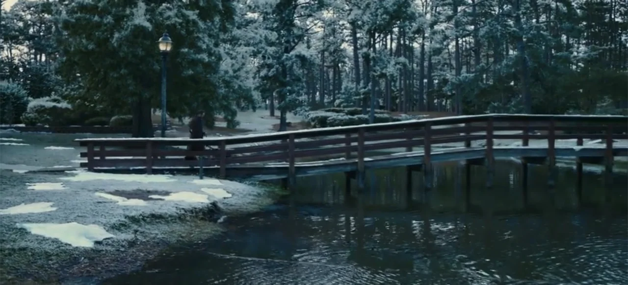 Merv (2025) movie still: wide — A snow-dusted park features a wooden footbridge crossing a dark pond in winter. A man in a dark coat walks across…