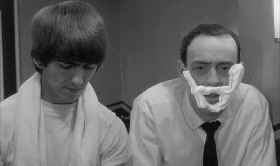 A Hard Days Night (1964) movie still: medium — A cramped bathroom space with plain walls and minimal visible fixtures. One man wears a white t-shirt…