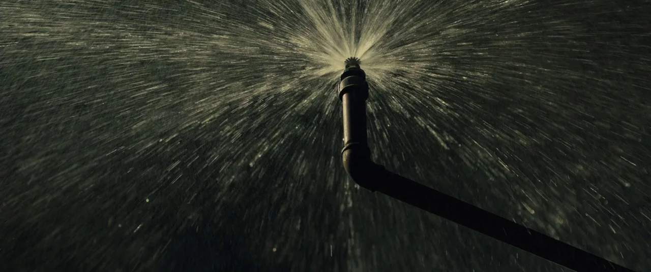Free Fire (2016) movie still: closeup — A dark outdoor space at night with a metal sprinkler nozzle at the center. High-pressure water shoots…
