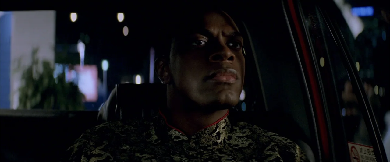 Rush Hour 2 (2001) movie still: medium — A man sits inside a dark vehicle at night with out-of-focus city lights visible outside. He wears a dark…
