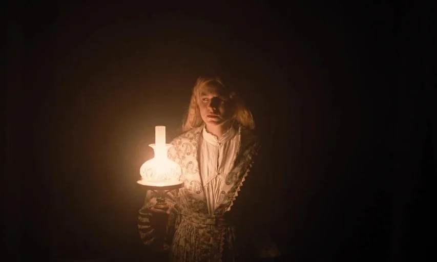 Nosferatu (2024) movie still: medium — A dark, unlit interior space surrounds a young woman dressed in period clothing. She holds a glowing oil lamp…