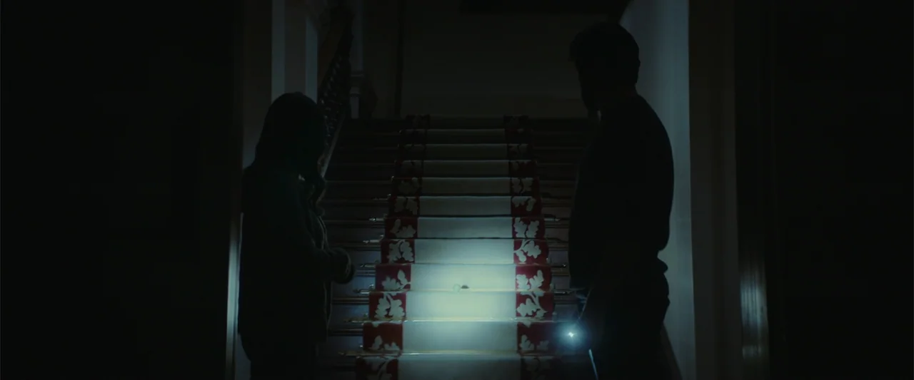 The Cellar (2022) movie still: medium — A dark residential interior features a central staircase with a patterned white and red runner. A man and a…
