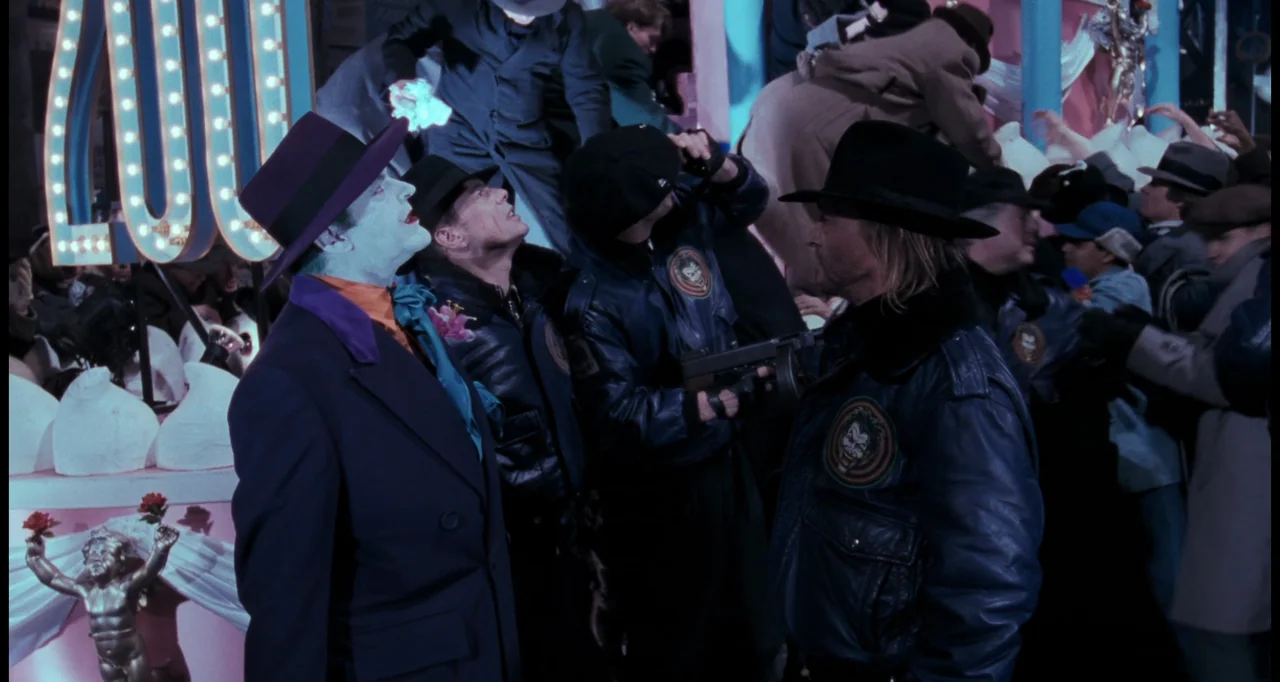 Batman (1989) movie still: medium — A night exterior set featuring a brightly lit parade float decorated with ornate sculptures. A man with white…