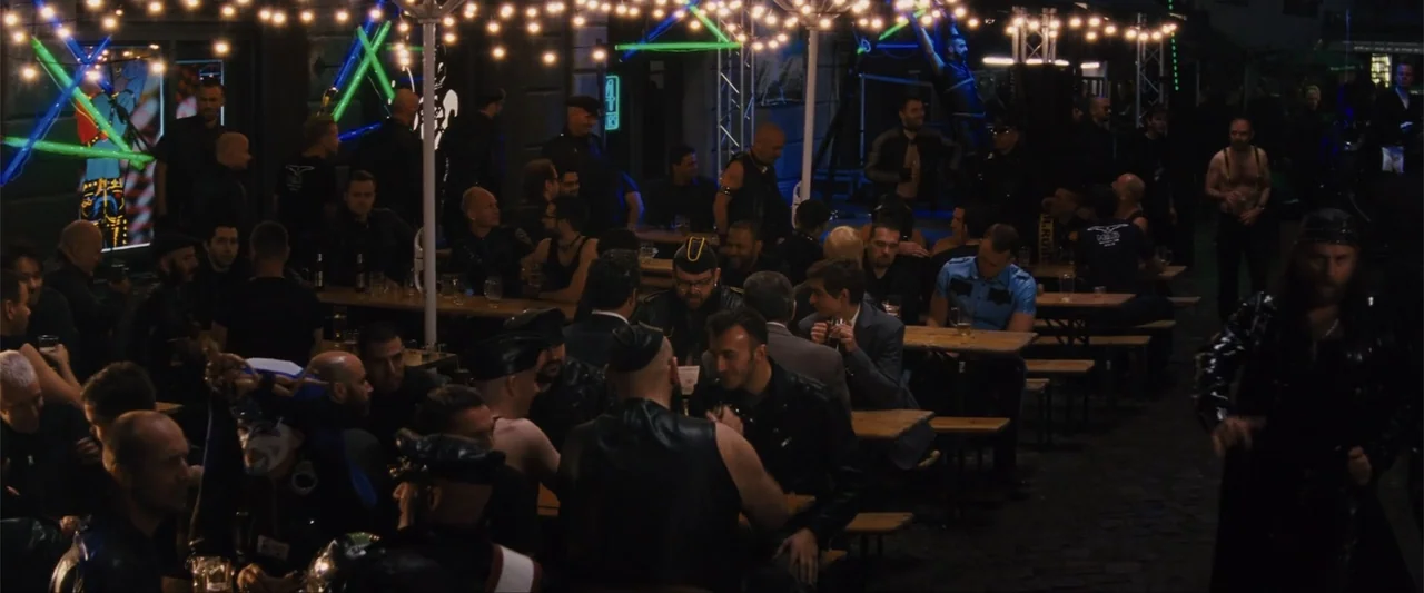 Unfinished Business (2015) movie still: wide — An outdoor beer garden at night with multiple wooden tables and string lighting overhead. A large…