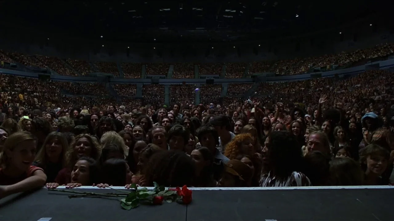 Almost Famous (2000) movie still: wide — A dark, massive concert arena is filled with a dense, dimly lit crowd of people. Two red roses lie…