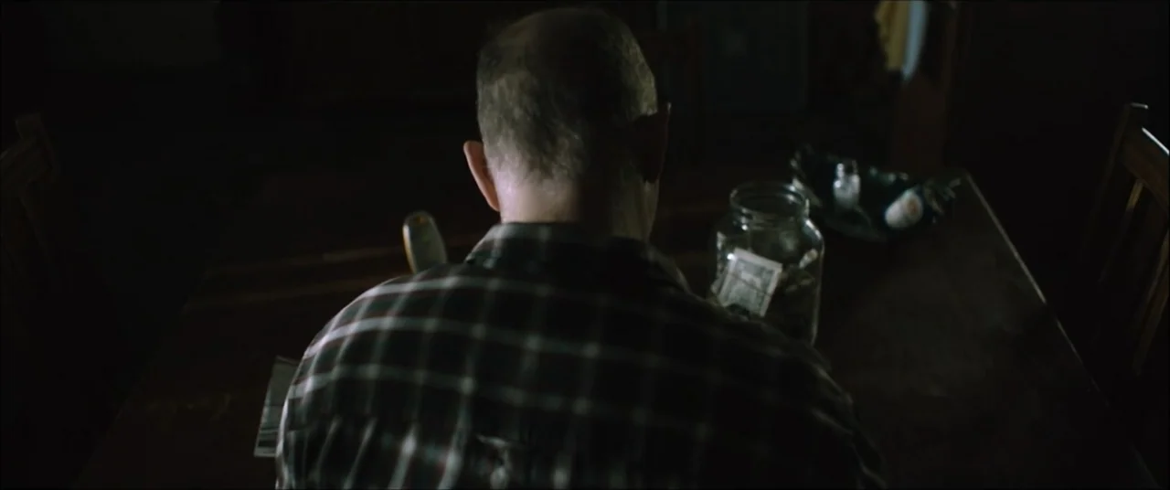 I'll Take Your Dead (2018) movie still: medium — A dark dining room with a man sitting at a wooden table at night. The man in a plaid shirt looks…