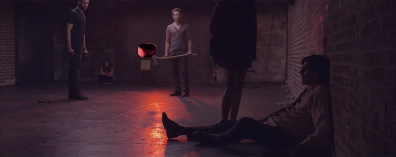 El Último Juego (2025) movie still: wide — A dark industrial basement with exposed brick walls and a concrete floor. A group of people stands around…