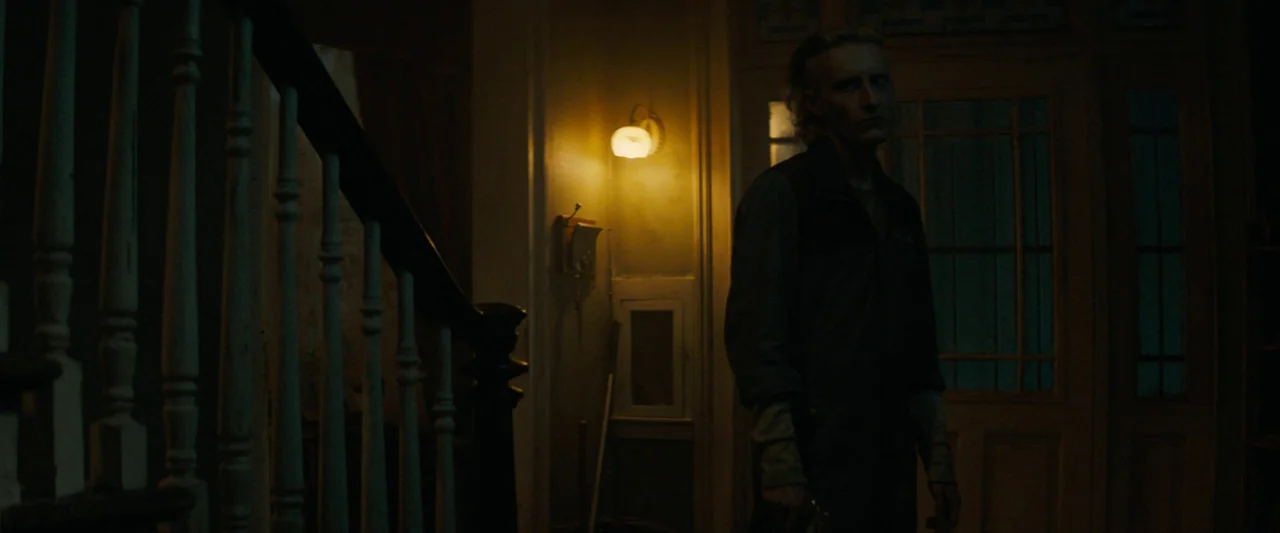 Don’t Breathe 2 (2021) movie still: medium — A dim building interior features a dark wooden staircase next to a wall light. A thin young man stands…