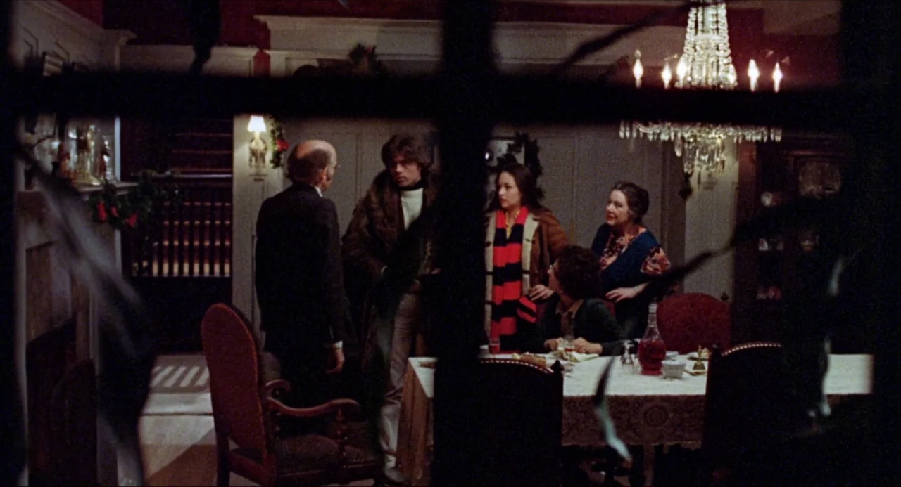 Black Christmas (1974) movie still: wide — A dimly lit 1970s dining room features wood paneling and a crystal chandelier. Five people stand or sit…