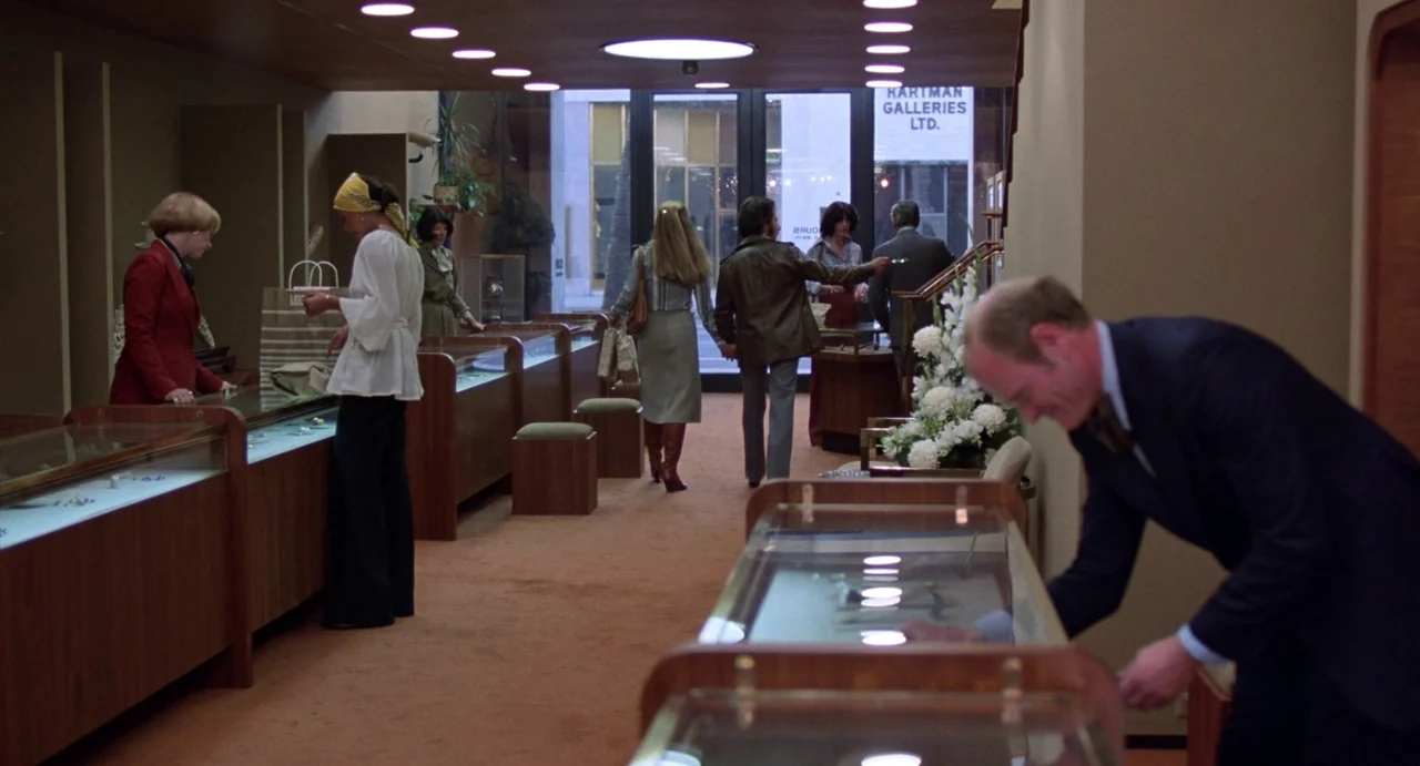 Straight Time (1978) movie still: wide — A well-lit 1970s jewelry store features wooden display cases and orange carpet flooring. Several patrons…