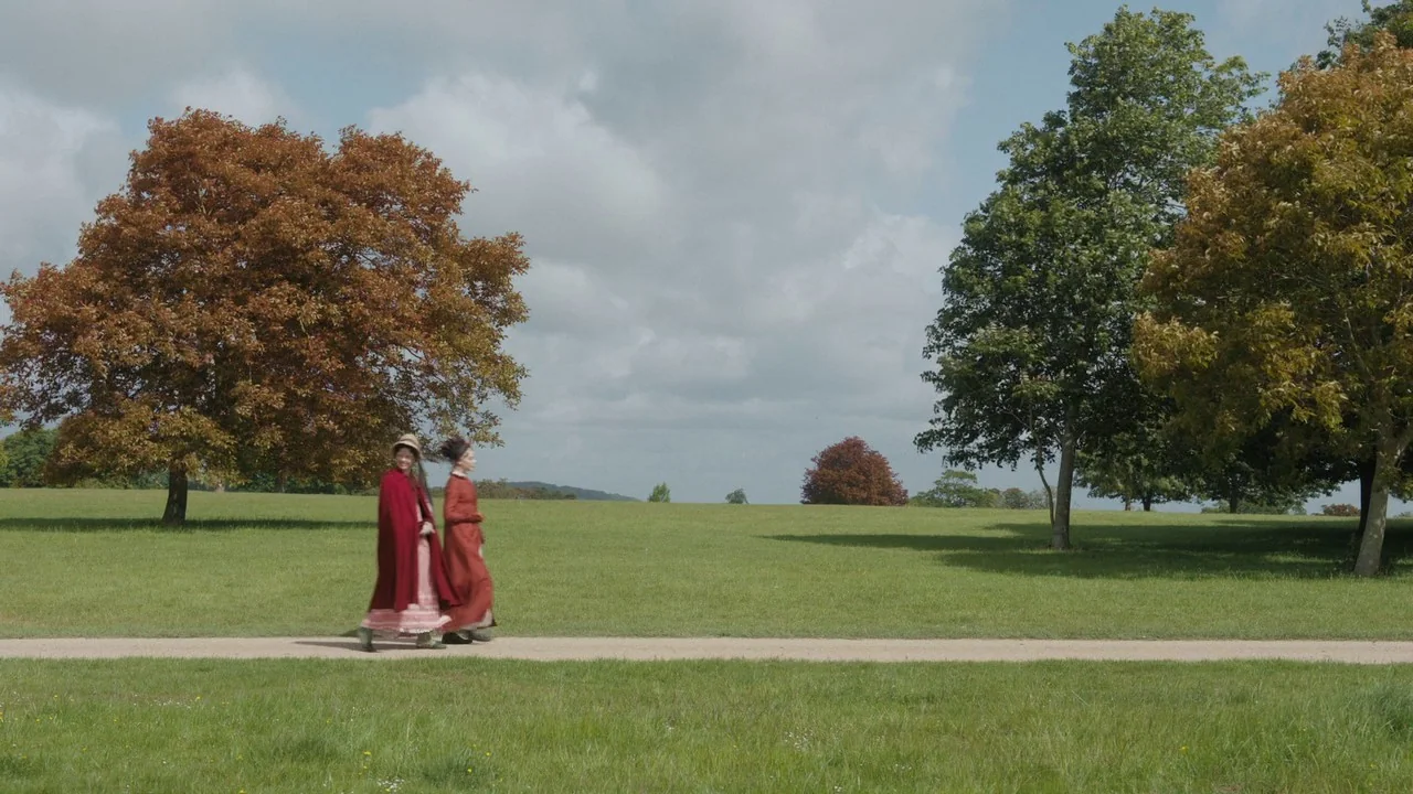 Emma. (2020) movie still: wide — A wide expanse of green parkland features large deciduous trees under a cloudy sky. Two women dressed in…