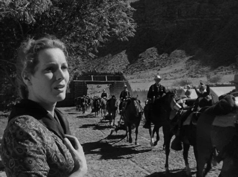 Rio Grande (1950) movie still: medium — A dusty outdoor cavalry post features wooden fortifications and horses held by mounted soldiers. A woman in…