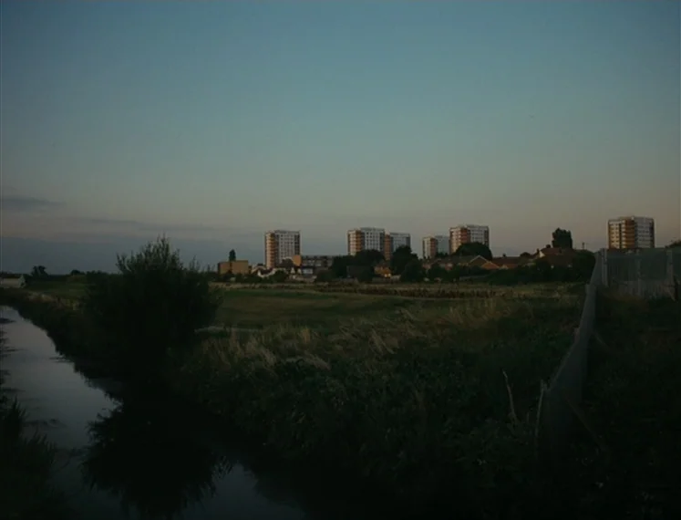 Fish Tank (2009) movie still: establishing — A dark canal reflects the sky next to a grassy field with distant apartment buildings. The scene…